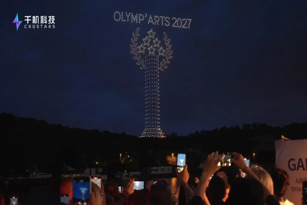 Crostars drone show forming OLYMP'ARTS 2027 text and Olympic torch shape over the Seine River at night, Paris July 2025 — audience photographing the display along the riverbank