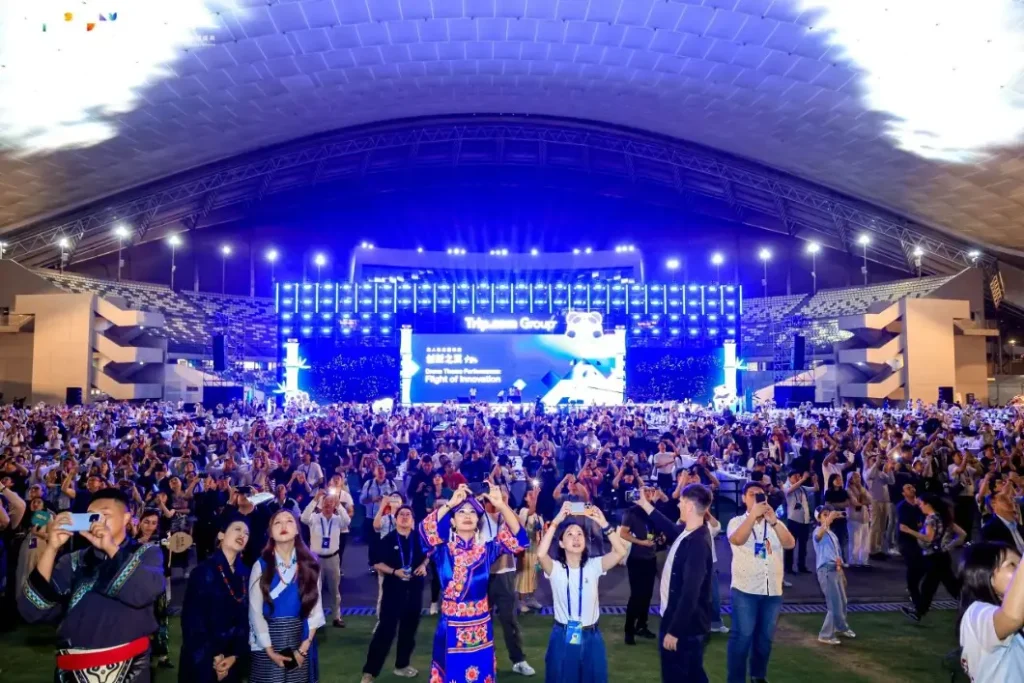 Trip.com Group Global Partner Conference 2025 indoor venue — thousands of attendees photographing the drone show, LED screen shows Flight of Innovation drone performance theme, traditional costume attendees visible